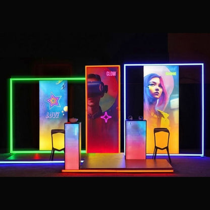 RGB lit display set at a night event: RGB Rotating Display frames lit green and blue with backlit panels inside, a freestanding backlit SEG wall and two lit plinths